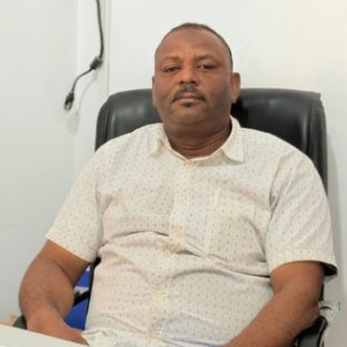 Dr. Mustafa Sharfi, wearing a light patterned short-sleeve shirt and seated in a black leather office chair in front of a white wall