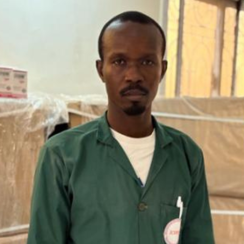 Mr. Daoissem Flaven, wearing a green work coat in a warehouse with stacked boxes behind him